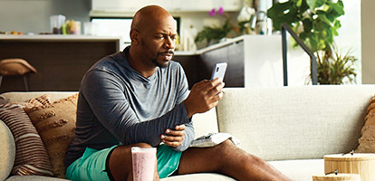 A man in shorts sitting on a couch with an icepack on his left leg while scheduling an appointment on his phone.
