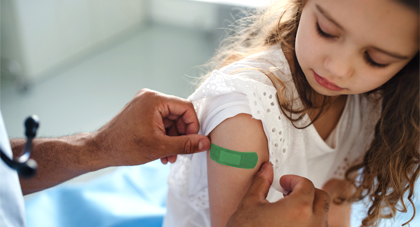 A young girl frowing while looking at her arm as a medical professional's hands place a band aid on her upper arm