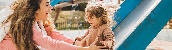 A laughing woman catching a smiling child at the bottom of a playground slide