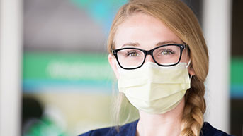 A nurse with glasses, wearing mask looking at the camera.