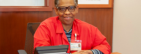 An AHN volunteer wearing their red AHN volunteer coat sitting at a desk and smiling at the camera.