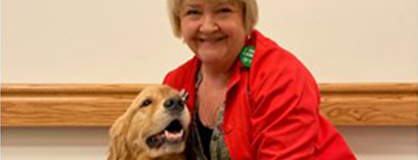 An AHN volunteer wearing their red AHN volunteer coat smiling and petting her therapy dog.
