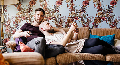 A young man laying on a tan couch next to his partner, using his smartphone to message his doctor on the MyChart app.