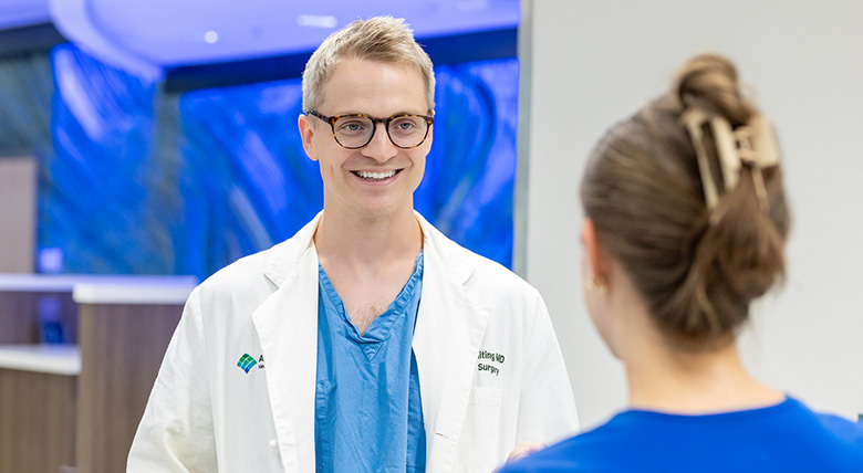 A smiling white medical professional in a white coat and glasses talks to someone in a blue scrubs.