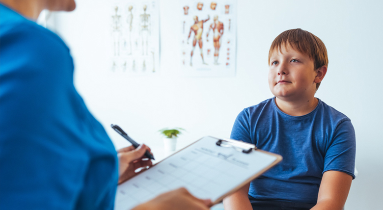 A medical professional in blue scrubs writes on a clipboard while a young boy looks up.