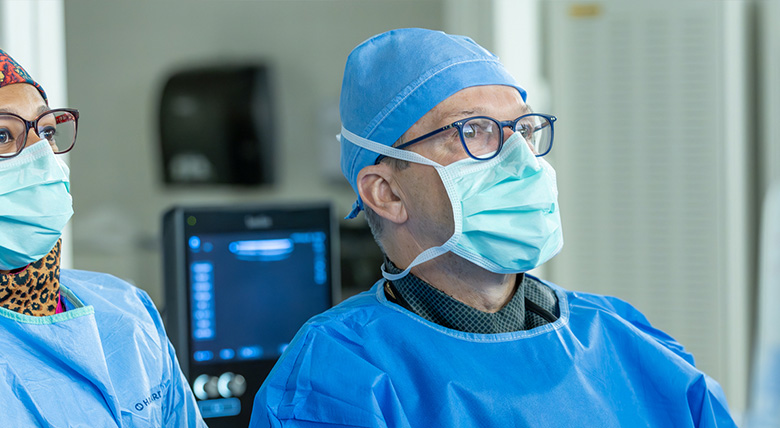 Two medical professionals in blue gowns and masks look focused in a hospital room.
