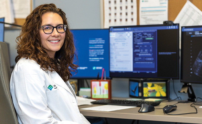A smiling woman in a white coat works at her computer in an office at the hospital.