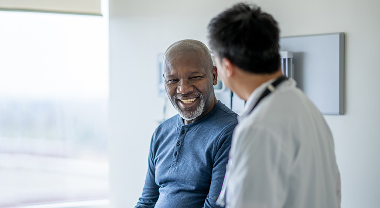 A smiling Black man talks to a medical professional wearing a white coat in a bright room.