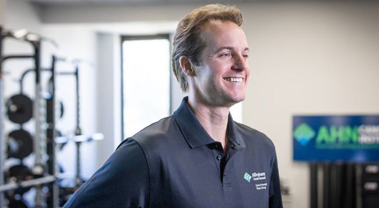 A smiling white medical professional in a black polo shirt looks happily to the right in a gym.