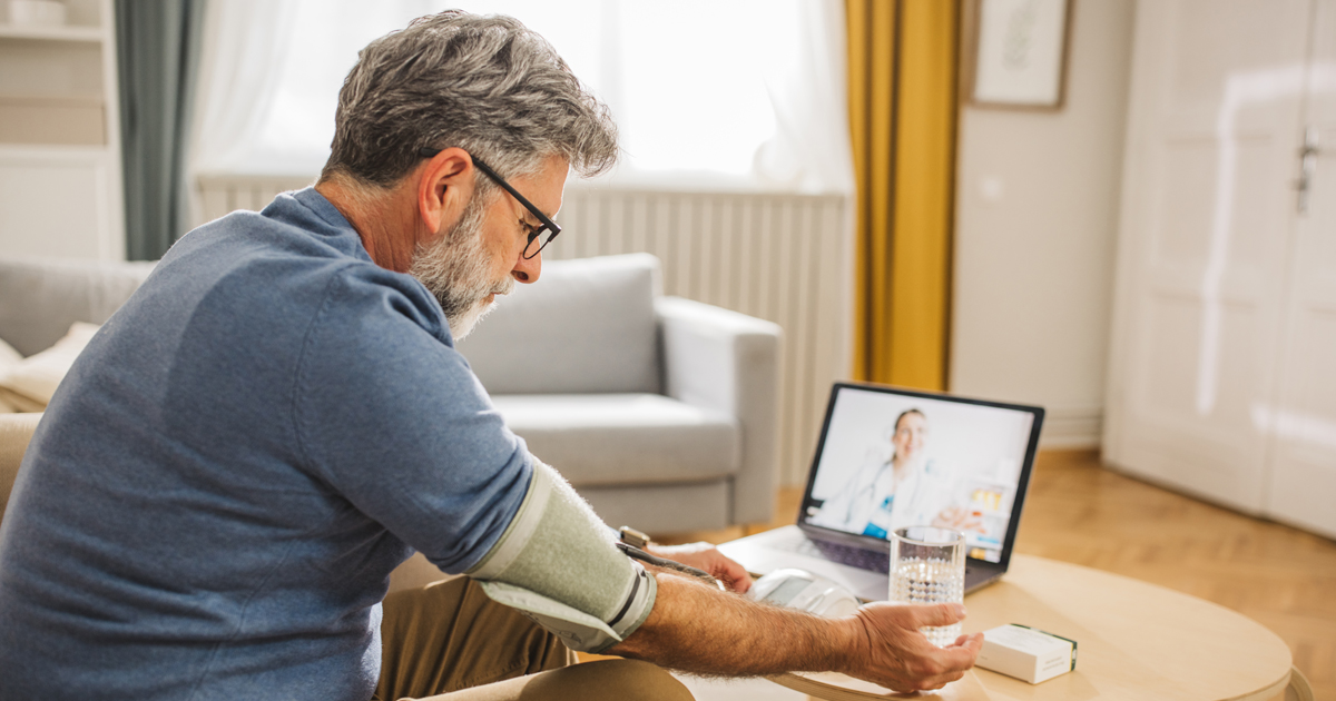 A white man wears a blood pressure cuff while video chatting with a medical professional.