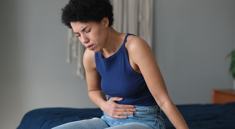A black woman in a blue tank top holds her stomach, looking like she is in pain.