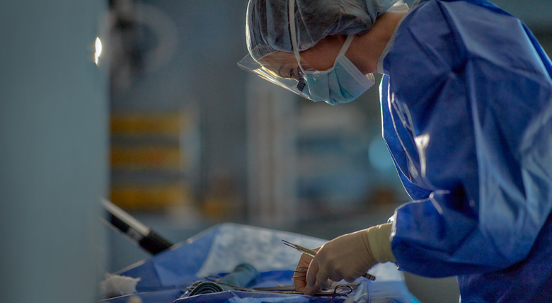 A medical professional in blue scrubs, a mask, and a face shield is working in an operating room.