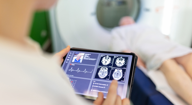 A medical professional in a white coat looks at brain scans on a tablet while a patient is in an MRI machine.