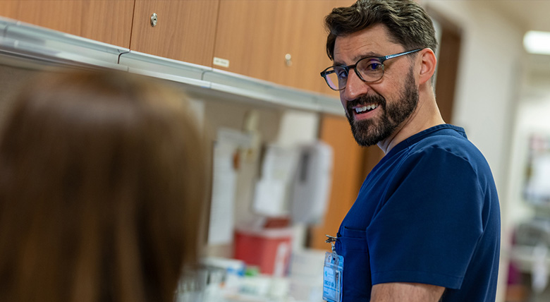 A White medical professional with glasses and a beard wears blue scrubs and smiles.