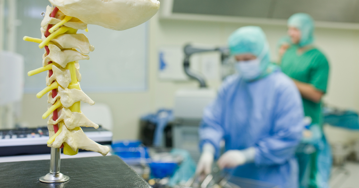 A model of a spine stands in a surgical room with two medical professionals in scrubs.