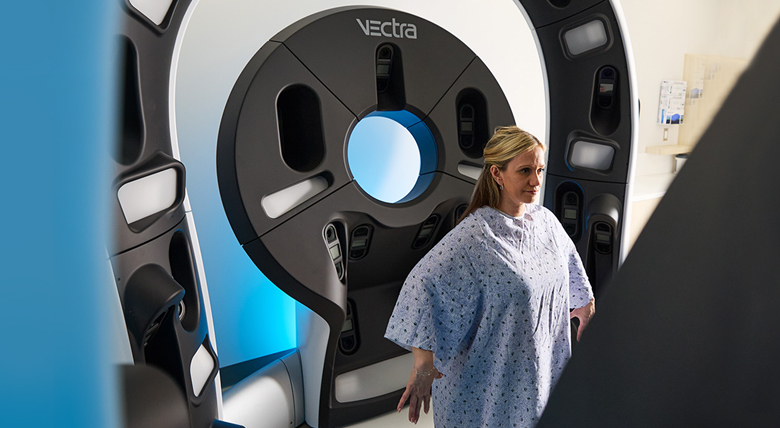 A blonde woman in a hospital gown stands before a VECTRA imaging machine.