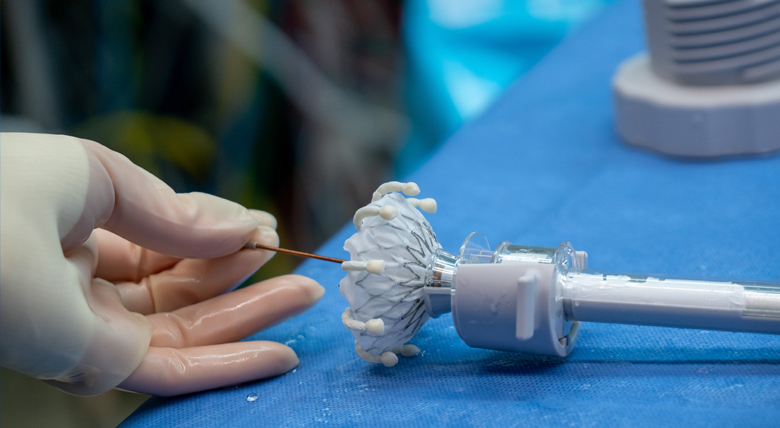 A medical professional in white gloves holds a small wire near a white and gray heart valve device on a blue cloth.