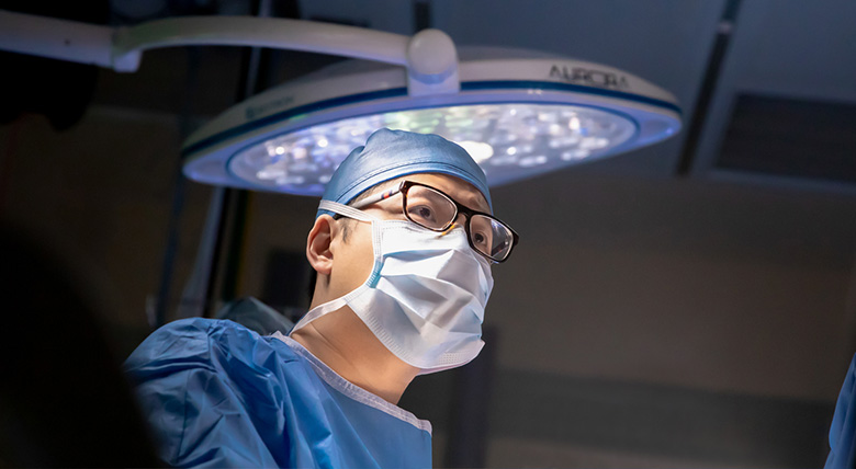 A surgeon in blue scrubs and a mask looks focused while working in an operating room with a bright light above.