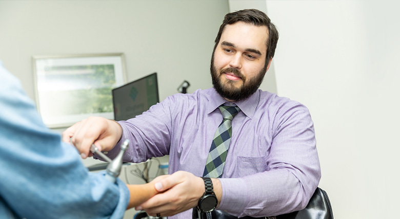 A White medical professional with a beard, wearing a purple shirt and tie, checks a patient's arm in an office.