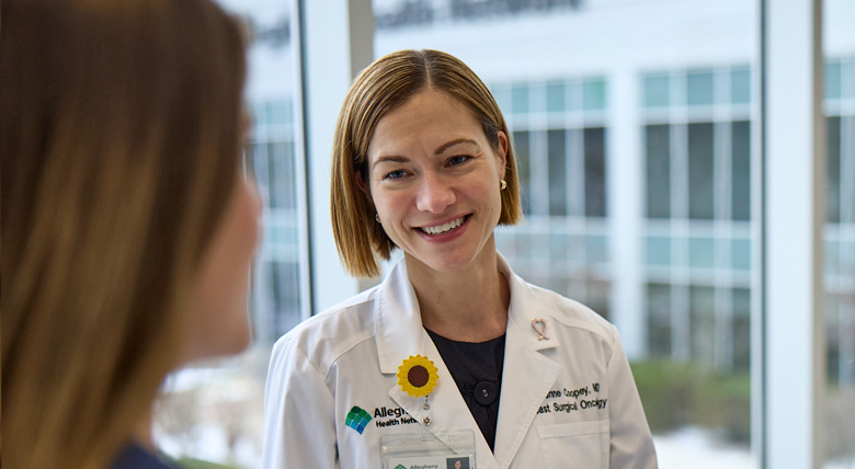 A smiling white female medical professional in a white lab coat talks to a patient near a bright window.