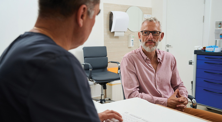 A medical professional wearing blue scrubs types on a keyboard as they talk to a white man.