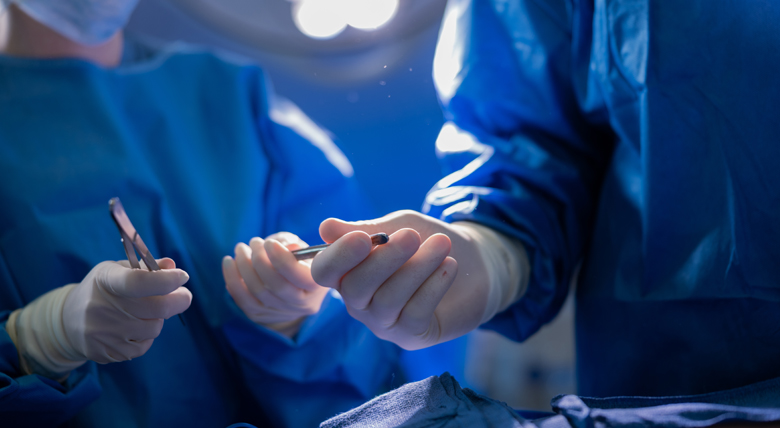 Two medical professionals in blue scrubs and white gloves hold surgical tools.