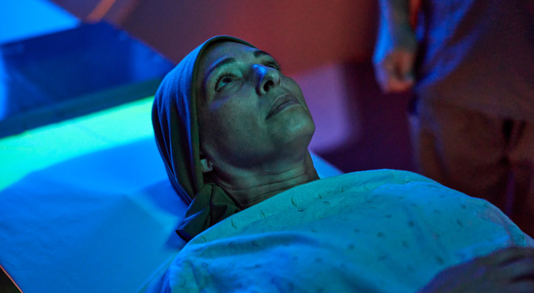 A white woman in a headscarf lies on a table, looking up with hope in a room lit with blue light for cancer care.