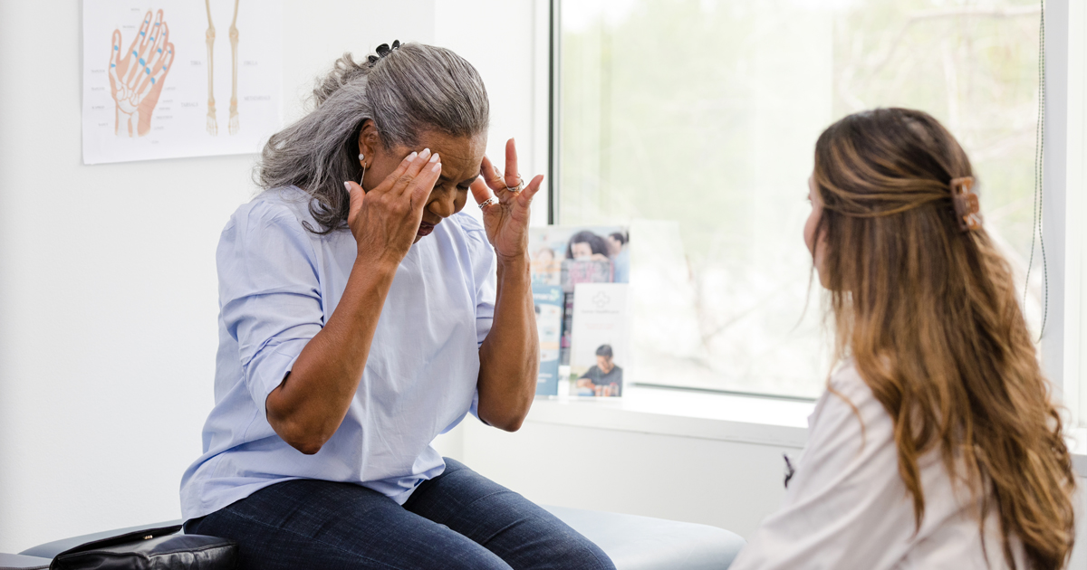 A Black woman patient with a headache meets with a medical professional in a white coat by a window.