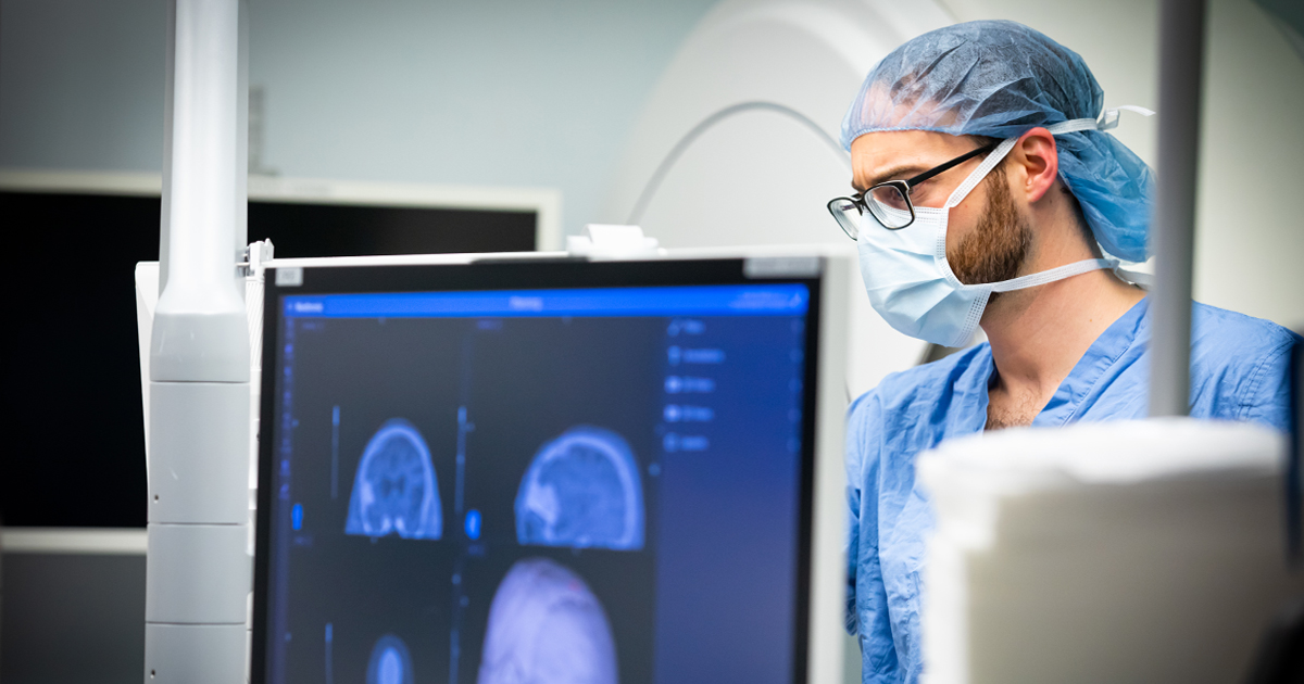 A White medical professional in blue scrubs, glasses, and a mask looks at a brain scan on a screen.