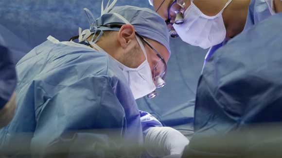 Two surgical residents wearing surgical gowns, PPE and surgical loupes, leaning in to perform a procedure