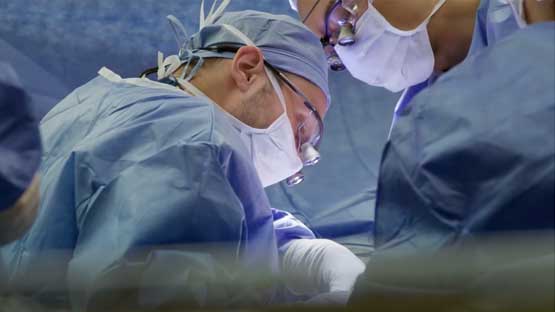 Two surgical residents wearing surgical gowns, PPE and surgical loupes, leaning in to perform a procedure