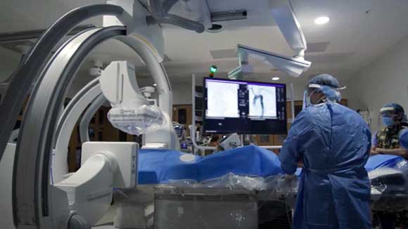 A diganostic radiology resident performs a procedure in a surgical suite with large monitors.