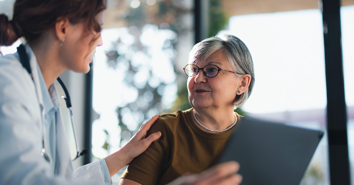 A medical professional in a white coat talks to an older woman, putting a hand on her shoulder.