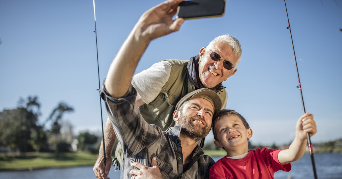Three generations of men smile while taking a selfie outdoors with fishing rods.