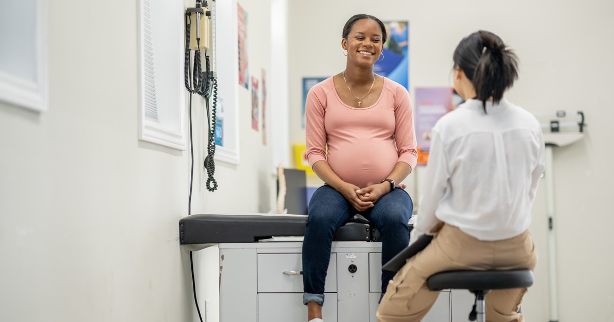 A smiling Black pregnant woman talks to a medical professional in a white coat.