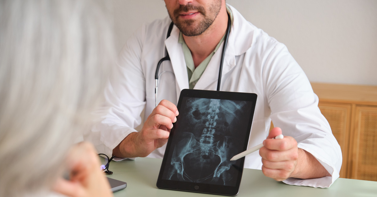A medical professional in a white coat shows a patient a spine x-ray on a tablet.