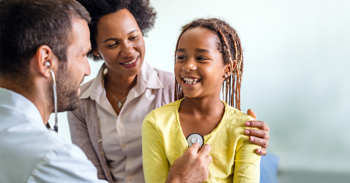 A medical professional examines a smiling girl using a stethoscope, as her mom watches over her.