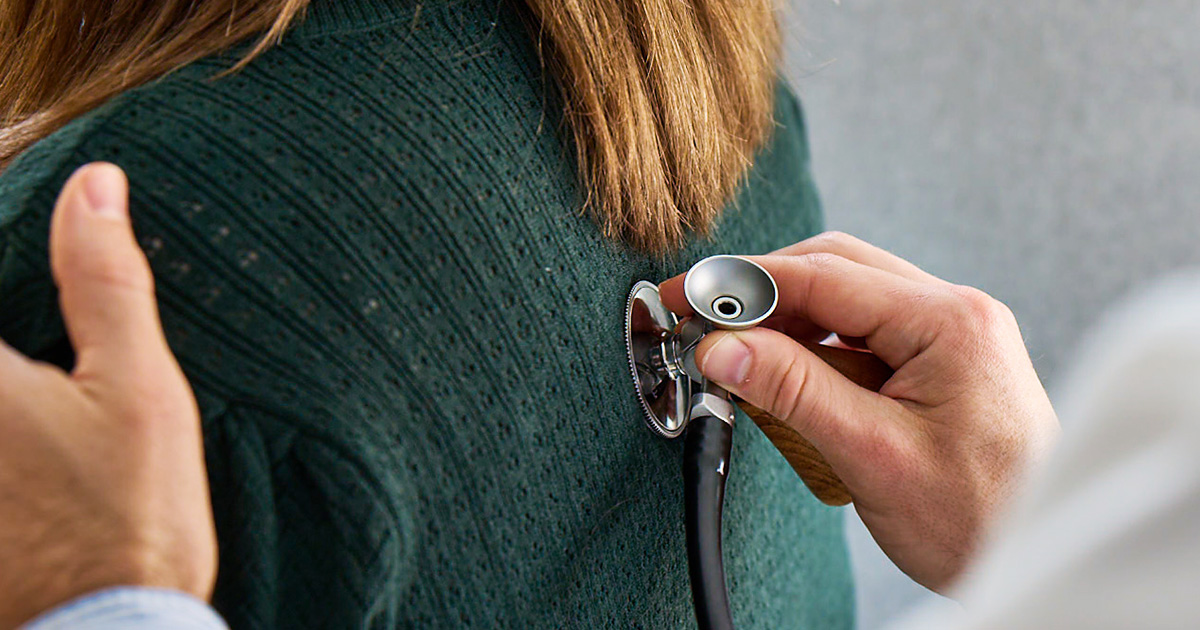 A medical professional in a white coat uses a stethoscope to listen to a person's back.