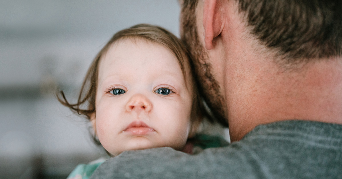 A small baby with blue eyes looks out from over a man's shoulder.