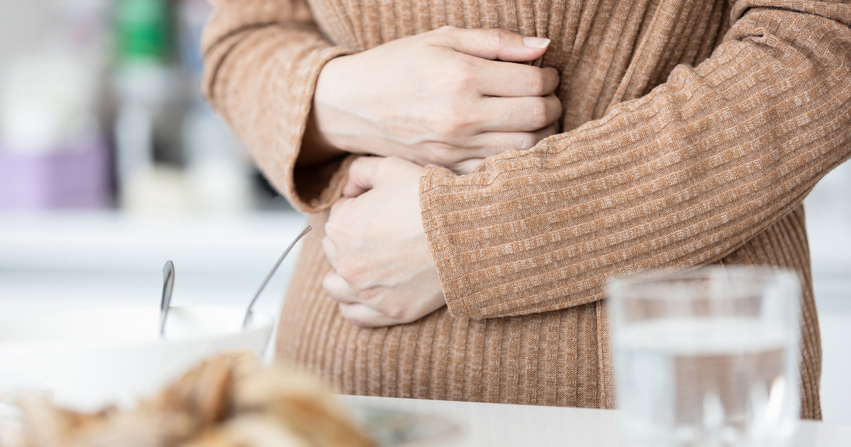A woman in a brown sweater holds her stomach, looking uncomfortable.