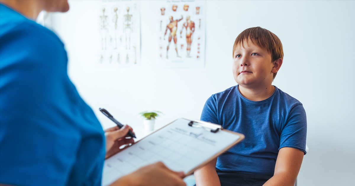 Medical professional in blue scrubs talks to a boy in a doctor's office.