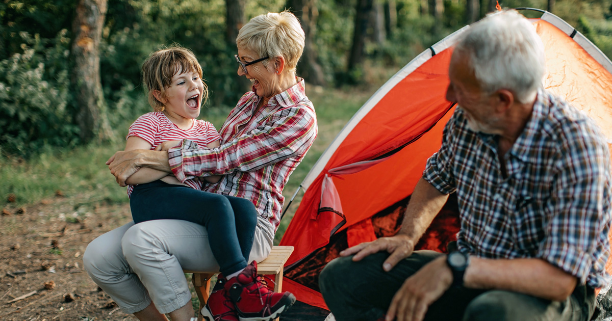 A grandma and grandpa laugh with their granddaughter while camping.