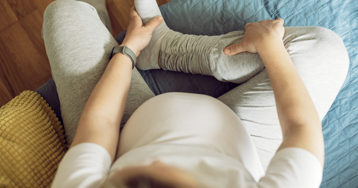 A pregnant person in light clothes sits and holds their lower leg.