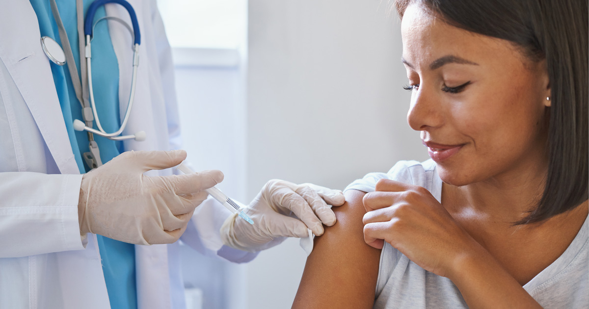 A medical professional in a white coat gives a Hispanic woman a shot in her arm.