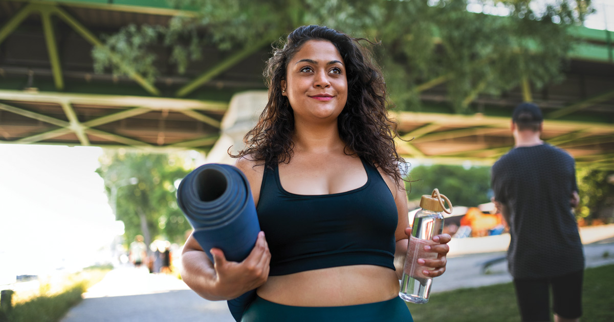 A smiling Indian woman holds a yoga mat and water bottle outside.