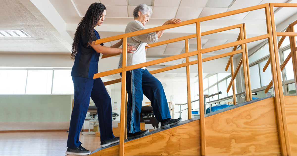 A medical professional in blue helps an older woman walk up a ramp.