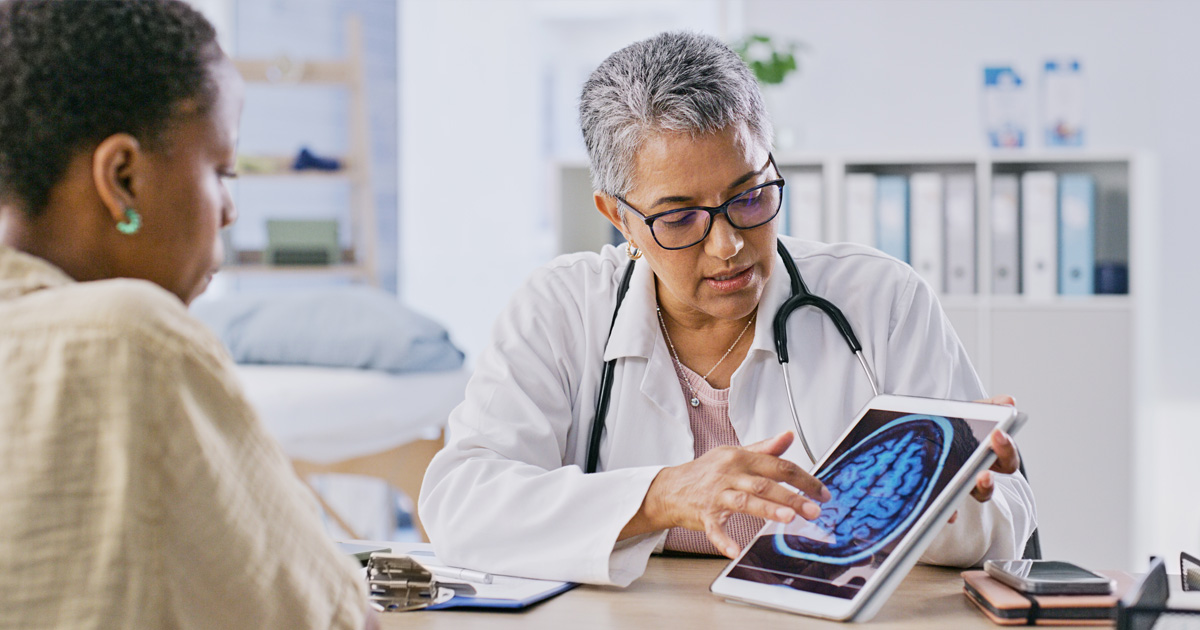 A medical professional in a white coat shows a brain scan on a tablet to a patient.