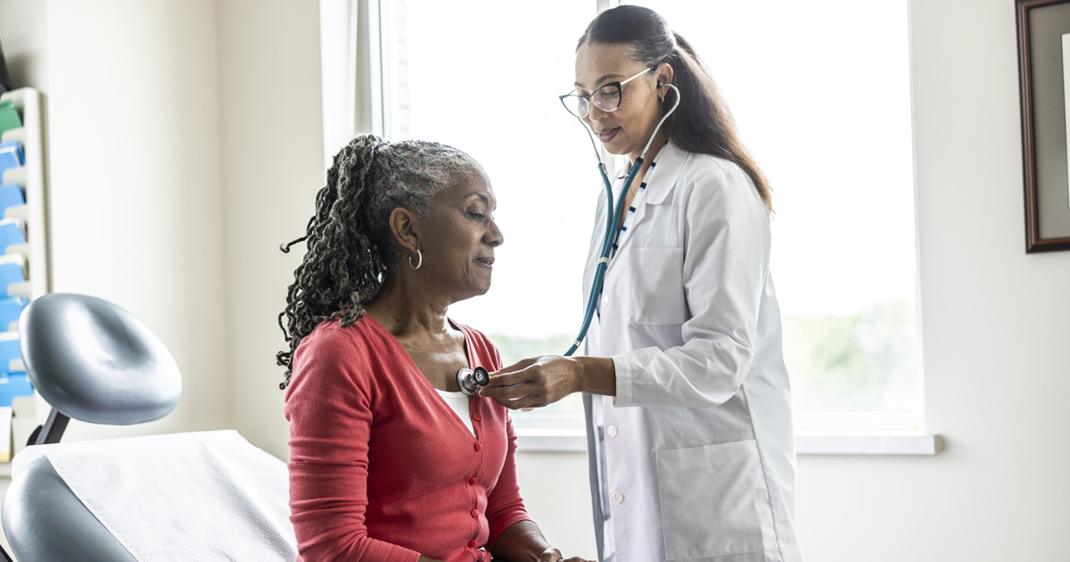 A Black medical professional in a white coat listens to a Black woman's chest with a stethoscope.