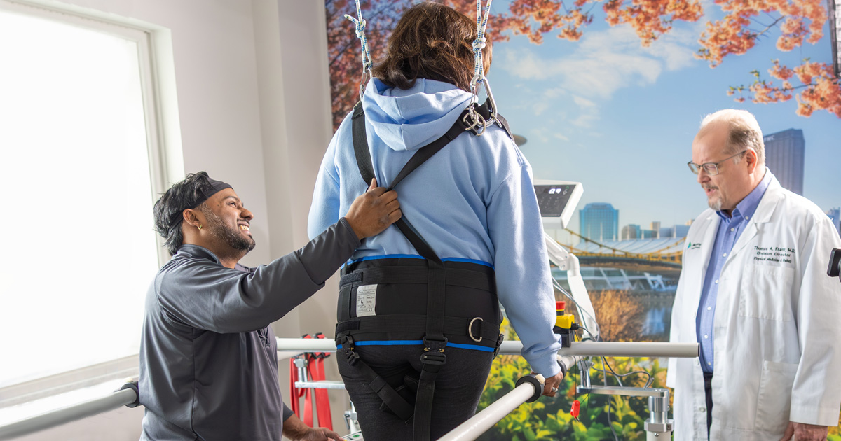 Two medical professionals help a woman in a harness walk.