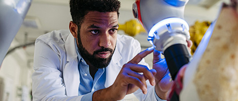 A black, male medical professional in a lab coat carefully examines a robotic arm with a blue light.